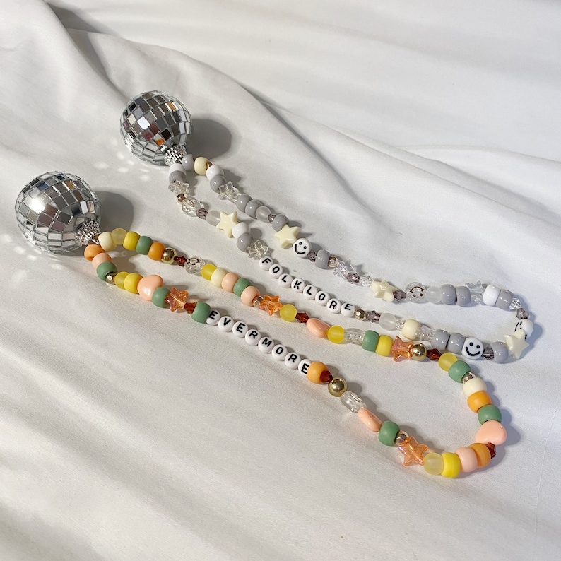 May include: Two beaded necklaces with silver disco ball charms. The necklaces are made with grey, green, yellow, and orange beads. One necklace has the word "folklore" spelled out in white beads and the other has the word "evermore" spelled out in white beads.