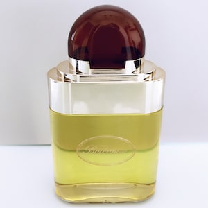 May include: A clear glass bottle with a gold cap and a brown dome-shaped top. The bottle contains a yellow liquid and has the word "Balmain" printed on the label.