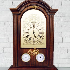 Puede incluir: Un reloj de pie de madera con una esfera dorada y marcadores de hora con números romanos. El reloj tiene dos diales más pequeños debajo de la esfera principal.