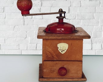 Puede incluir: Un molinillo de café vintage de madera con una tapa de metal roja y una manivela. El molinillo tiene un cajón para recoger los posos de café y una pequeña perilla roja en la parte delantera.