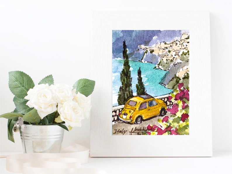 May include: Watercolor painting of a yellow Fiat car driving along a winding road overlooking a blue ocean and a small town on a cliffside. The painting is framed in a white frame and has the text "Italy" written at the bottom.