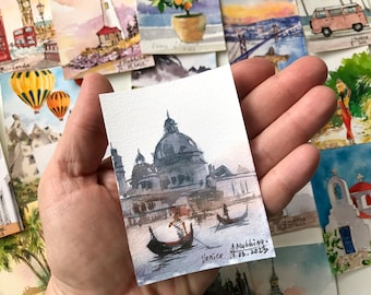May include: A watercolor painting of a building with a dome in Venice, Italy. The painting is on a small piece of paper and is held in a hand. The painting is signed "A. Mykhina, 08.08.2023" and "Venice".