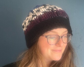 May include: A black and white knit beanie with a purple and blue stripe. The beanie is worn by a person with long blonde hair and glasses.