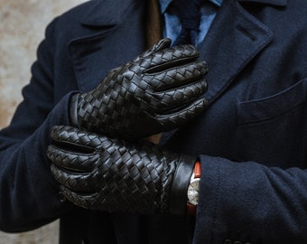 May include: A pair of black leather gloves with a woven pattern. The gloves are being held by a person wearing a dark blue coat.