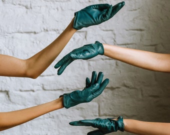 May include: Three hands wearing teal leather gloves are outstretched against a white brick wall background.