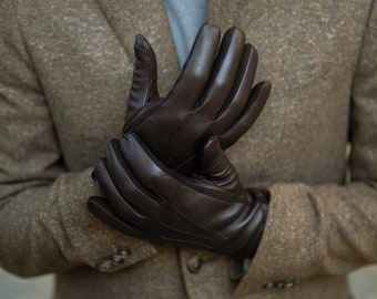 May include: A pair of brown leather gloves with a textured surface. The gloves are folded over each other and are being held by a person.