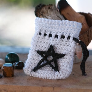 Peut inclure: Une pochette blanche en crochet avec un motif de pentagramme noir et un cordon de serrage noir. La pochette est pos&eacute;e sur une surface en bois avec divers cristaux et pierres.