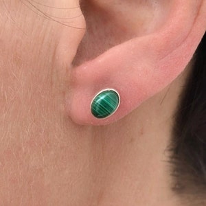 Könnte beinhalten: Nahaufnahme eines Ohrs mit einem Malachit-Ohrstecker. Der ovale Ohrring hat eine silberne Fassung und einen grünen, gestreiften Stein. Der Ohrring ist im Ohrläppchen eingesetzt.