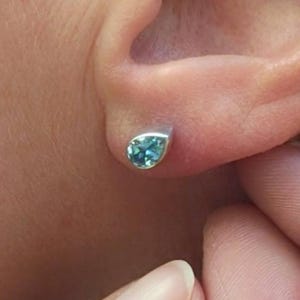 Op de afbeelding: Een close-up van een zilveren oorbel met een druppelvormige, lichtblauwe edelsteen. De oorbel is gezet in een eenvoudig, modern design en wordt op een oor gedragen. De afbeelding toont de oorbel in detail.