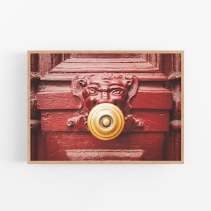 Puede incluir: Primer plano de una puerta de madera roja con un llamador de puerta de oro en forma de cara. El llamador de puerta tiene un botón redondo de oro en el centro.