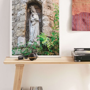 May include: A framed photograph of a white stone statue of the Virgin Mary and baby Jesus in a stone alcove with green foliage. The statue is set against a backdrop of a stone wall.