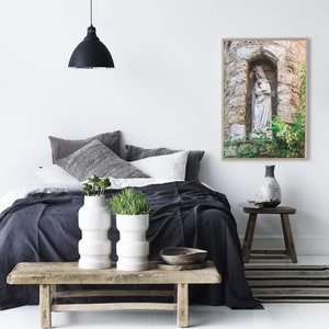 May include: A white statue of a woman holding a child in a stone alcove with green plants. The statue is in a framed photograph hanging on a white wall above a bed with a dark gray blanket and white pillows.