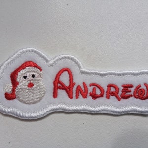 Pode incluir: Ornamento de Natal de feltro branco com um Papai Noel bordado em vermelho e o nome "ANDREW" em bordado vermelho.
