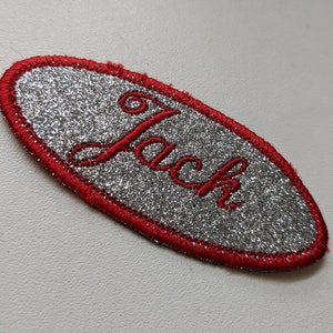 May include: Silver glitter oval name tag with a red stitched border. The name "Jack" is embroidered in red.