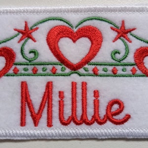 May include: White felt patch with a green and red embroidered design. The design features three red hearts with white centres, two red stars, and green swirls. The name "Millie" is embroidered in red below the design.