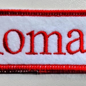 May include: Embroidered patch featuring the name "Roman" in red thread on a white rectangular base. The patch is framed by a red, intricately stitched border, designed for attachment to garments or bags.
