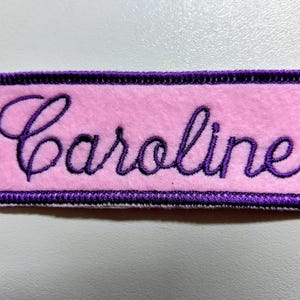 May include: A rectangular, pink embroidered patch with the name "Caroline" in purple cursive script. The patch has a purple border and is likely designed to be sewn onto clothing or accessories.