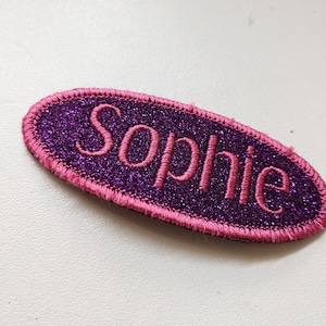 May include: Purple glitter oval-shaped patch with pink stitching and the name "Sophie" embroidered in pink.