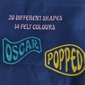 May include: A dark blue felt background featuring the embroidered text 20 DIFFERENT SHAPES 14 FELT COLOURS in light pink. Two embroidered patches are present: one teal wavy banner with the word OSCAR and one yellow oval with the word POPPED.