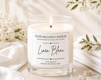 May include: A clear glass jar candle with a lit wick and white wax. The label reads: CUSTOM LOVE CANDLES, HAND-POURED NATURAL SOY CANDLE, Linen Blanc, 8 OZ (226.8 G), MADE IN PENNSYLVANIA. The candle is positioned among white fabric and flowers.