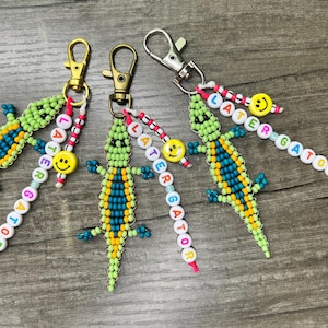 May include: Four beaded alligator keychains with gold and silver clasps. The alligators are green, blue, and yellow. Each keychain has a beaded word that says "LATER GATOR" and a yellow smiley face bead.