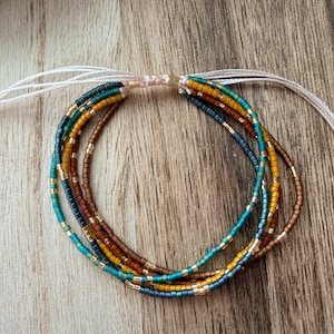 May include: A multi-strand bracelet featuring small seed beads in shades of teal, mustard yellow, and brown, accented with metallic gold beads. The strands are connected by a light-colored cord with adjustable sliding knots and gold-toned end caps.