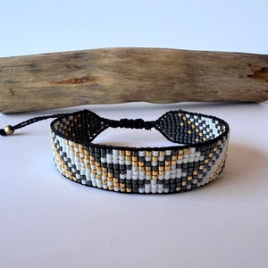 May include: Woven bracelet with a geometric pattern in black, white, and gold beads. The bracelet has a black adjustable cord closure and a small gold bead accent. The background includes a piece of driftwood.