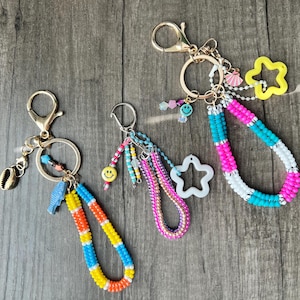 以下が含まれることがあります： ゴールドトーンの金具が付いた、ビーズのキーホルダー3つ。1つは貝殻のチャーム、もう1つはスマイリーフェイスのチャーム、もう1つは星のチャームが付いています。ビーズのストラップは、オレンジ、黄色、青、ピンク、白など、さまざまな色があります。
