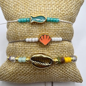 May include: Three beaded bracelets with ocean-themed charms rest on a burlap pillow. The top bracelet features a teal fish charm and turquoise beads. The middle bracelet has an orange seashell charm. The bottom bracelet has a gold seashell charm.