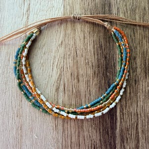 May include: A multi-strand bracelet featuring small cylindrical beads in shades of white, orange, blue, and green, interspersed with metallic gold-tone spacer beads. The strands are secured with a tan cord that features a sliding knot closure.