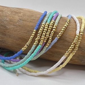 May include: A collection of colorful beaded bracelets in shades of blue, green, purple, and yellow. Each bracelet features gold-colored beads interspersed with the colorful beads. The bracelets are arranged on a piece of driftwood.