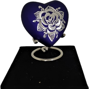 May include: A heart-shaped, navy blue urn with a silver rose design. The urn is displayed on a black velvet box with a gold clasp. The urn is supported by a silver metal stand.