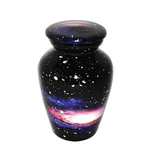 May include: A small black cremation urn with a galaxy design. The urn has a white lid and features a swirling nebula with pink, purple, and white colors.