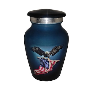 May include: A blue cremation urn with a black lid and silver trim. The urn features an image of a bald eagle with outstretched wings above an American flag. The urn is designed to hold the ashes of a loved one.