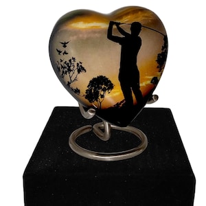Könnte beinhalten: Eine herzförmige Keramikurne mit einer Silhouette eines Golfspielers vor einem Sonnenuntergangshintergrund. Die Urne steht auf einem schwarzen Samtständer mit einem Metallfuß.