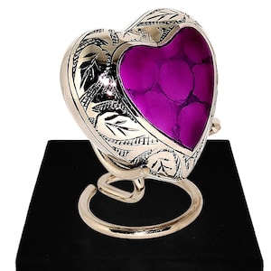 May include: A heart-shaped urn with a silver finish and engraved leaf design. The center features a vibrant purple stone-like material. It rests on a silver stand atop a black velvet box with a gold clasp. A memorial keepsake.