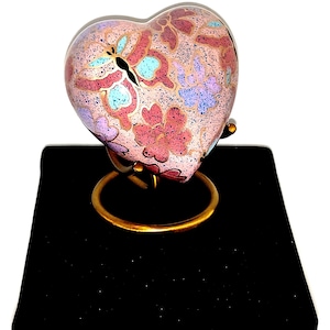 May include: A heart-shaped decorative urn with a floral and butterfly design in pink, blue, and purple. The urn sits on a gold-colored stand atop a black velvet box with a gold clasp.