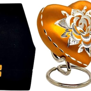May include: A gold heart-shaped cremation urn with a silver rose design. The urn is on a silver stand and comes with a black velvet box.