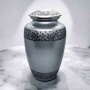 May include: Silver cremation urn with an ornate floral design around the top and bottom. The urn has a lid.