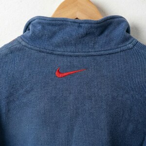 Puede incluir: Primer plano de una camiseta deportiva azul y roja. La camiseta tiene un cuello alto y detalles rojos en los hombros. Un logotipo Nike rojo est&aacute; bordado en el pecho. La tela tiene un aspecto texturizado y acanalado.