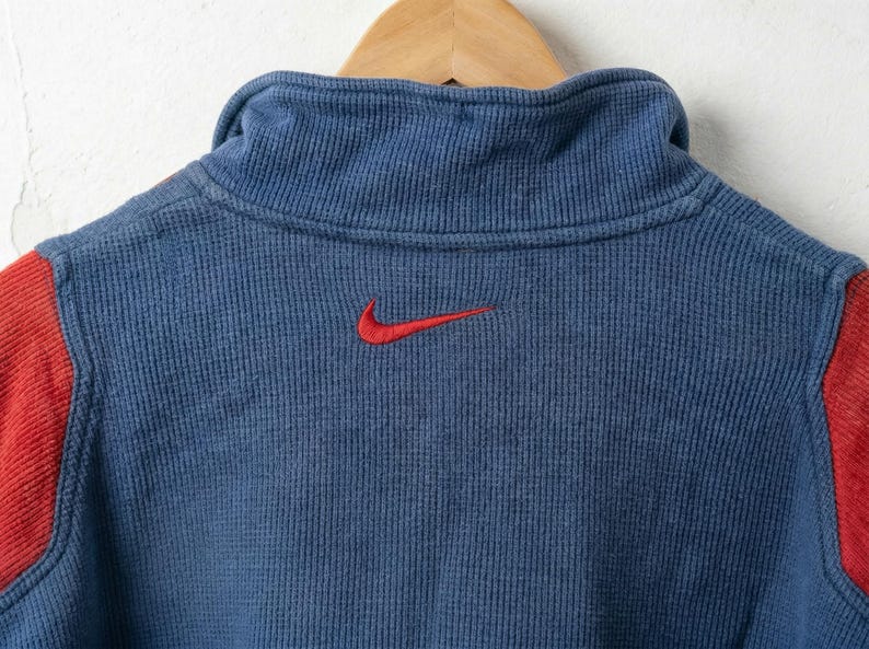 Puede incluir: Primer plano de una camiseta deportiva azul y roja. La camiseta tiene un cuello alto y detalles rojos en los hombros. Un logotipo Nike rojo est&aacute; bordado en el pecho. La tela tiene un aspecto texturizado y acanalado.
