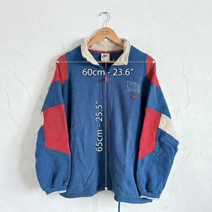 Puede incluir: Chaqueta con cremallera Nike azul, roja y crema. La chaqueta tiene cuello y el logo de Nike bordado en el pecho. La chaqueta mide 65 cm de largo y 60 cm de ancho.