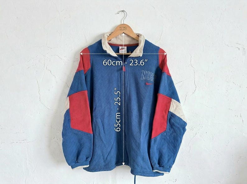 Puede incluir: Chaqueta con cremallera Nike azul, roja y crema. La chaqueta tiene cuello y el logo de Nike bordado en el pecho. La chaqueta mide 65 cm de largo y 60 cm de ancho.