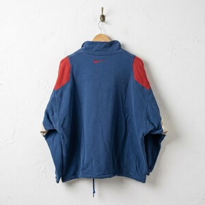 Puede incluir: Una sudadera Nike azul y roja colgada de una percha de madera. La sudadera tiene un cuello alto, parches rojos en los hombros y un peque&ntilde;o logotipo Nike rojo en la espalda. Las mangas tienen un acento color crema.
