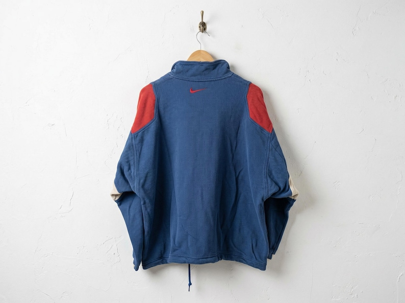 Puede incluir: Una sudadera Nike azul y roja colgada de una percha de madera. La sudadera tiene un cuello alto, parches rojos en los hombros y un peque&ntilde;o logotipo Nike rojo en la espalda. Las mangas tienen un acento color crema.