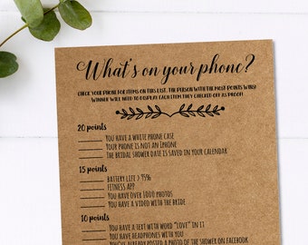 Può includere: Un biglietto di gioco in carta kraft con il testo "What's on your phone?" in corsivo nero. Il biglietto include un elenco di richieste di punteggio per un gioco di addio al nubilato. Rami verdi di eucalipto sullo sfondo.