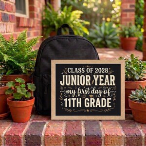 May include: A black backpack positioned behind a rectangular sign with a light wood frame. The sign features a black background with white text that reads: CLASS OF 2028 JUNIOR YEAR my first day of 11TH GRADE, decorated with small star and line icons.