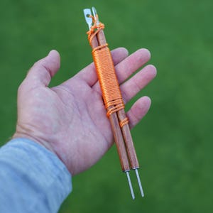 May include: Two parallel wooden rods joined by a bright orange cord wrapped tightly around the center. Each rod features a metallic tip with a small hole at the top end and a thin, threaded metal screw protruding from the bottom end.