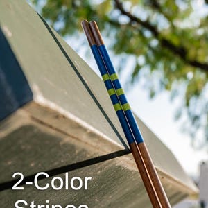 May include: Two wooden chopsticks with a brown base and blue and green striped design. The chopsticks are leaning against a green surface. The text "2-Color Stripes" is visible.