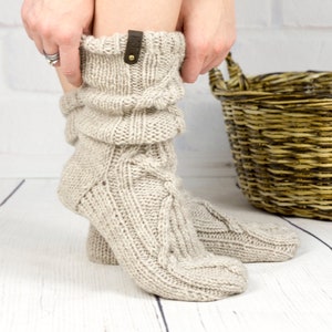 May include: A pair of beige knitted slouch socks with a brown leather tag. The socks are folded down at the top and have a cable knit pattern.